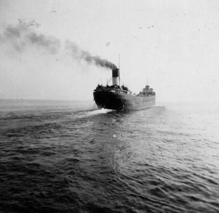 Black-and-white photograph of a large steamship moving across open water, trailing dark smoke and a broad wake.