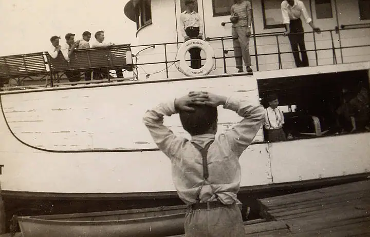 A child in a sailor-style outfit faces a passenger vessel from a dock while people look down from its decks.