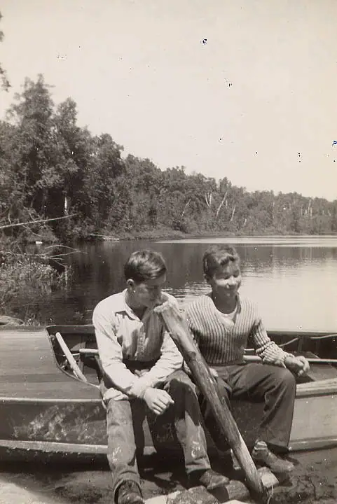 Two people sit in a small wooden boat beside a wooded body of water.