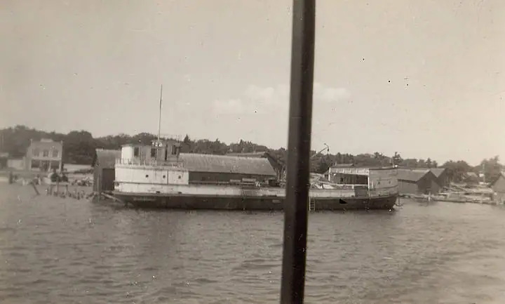 Black-and-white photograph of a long, low ferry beside waterfront buildings, partly obscured by a foreground pole.