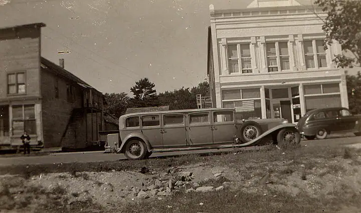 A long vintage automobile and another car sit beside a rough roadside in front of two wooden buildings.