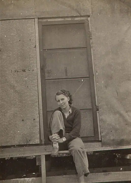 Black-and-white portrait of a woman seated on a wooden bench in front of a paneled door.