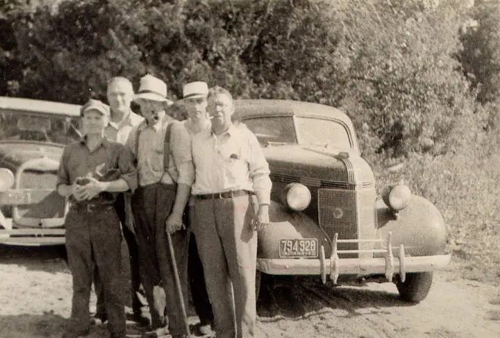 Five people stand beside two vintage automobiles in a wooded outdoor setting.