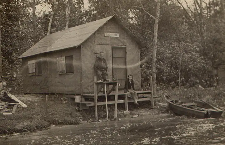 A weathered wooden cabin marked “SUNNYSIDE” stands beside the water, with two people and a small boat nearby.