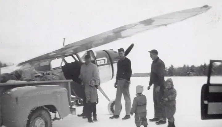 Six people stand beside a small airplane and pickup truck in a snowy open field.