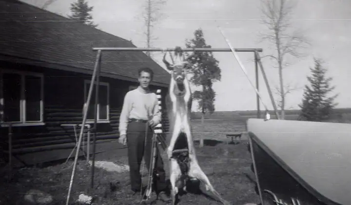 A person stands beside a hanging deer carcass outside a log building, with a boat hull at right.