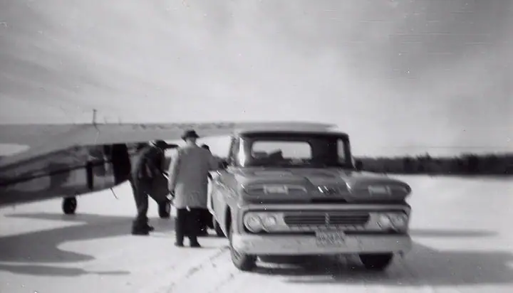 A vintage automobile and people stand beside a small airplane on a snow-covered field.