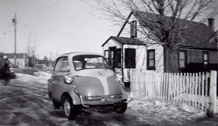 A small rounded car sits on a snowy residential street beside a wooden house and picket fence.