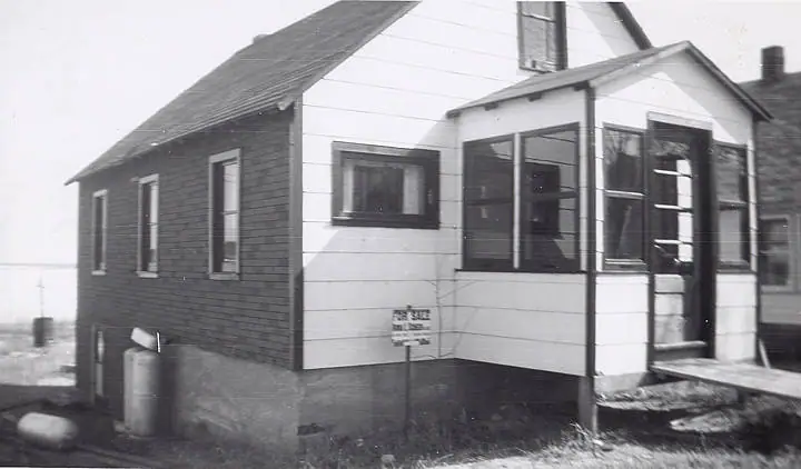 Black-and-white photograph of a small house with a screened porch and a FOR SALE sign.