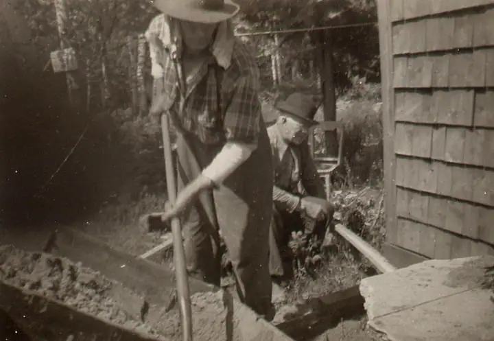 Two men work with hand tools beside a shingled wooden building.