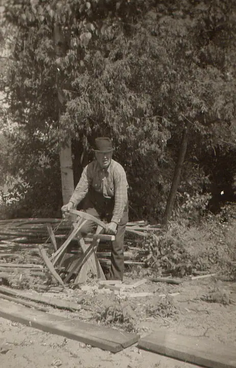 A man in a hat and overalls uses a bucksaw among cut wood in a wooded clearing.
