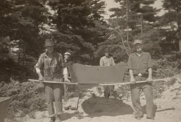 Four people handle a homemade wooden apparatus in a sandy clearing surrounded by trees.