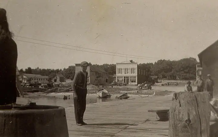 Black-and-white waterfront photograph with a man standing on a wooden dock among small boats and buildings.