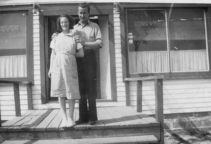 A man and woman stand together on a wooden porch outside a building with curtained windows.