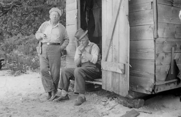 Black-and-white photograph of two men beside an open wooden shed, with one seated on the entrance step.