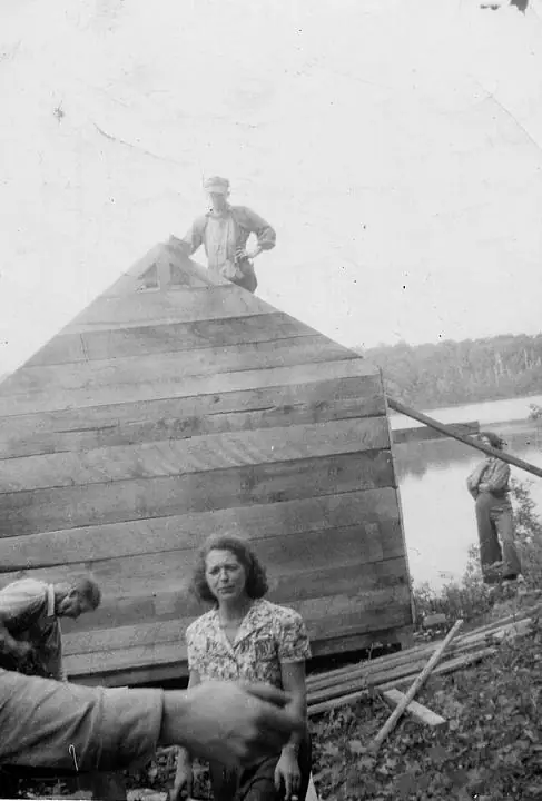 People work around a large wooden hull or structure beside the water.