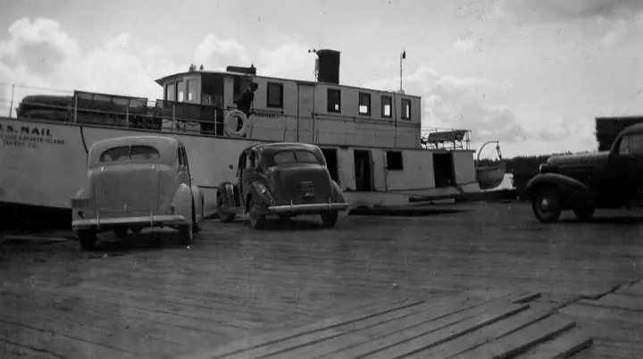 A ferry beside a wooden pier, with three vintage automobiles in the foreground.