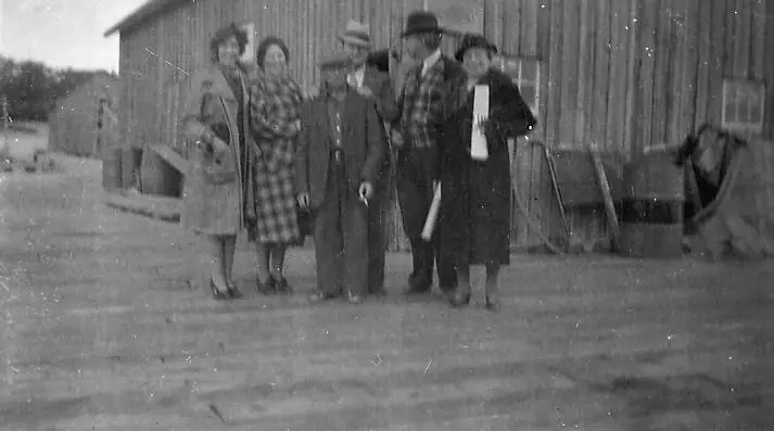 Six people pose outdoors beside a large wooden building in a black-and-white photograph.