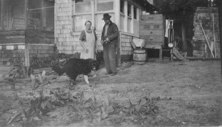 Black-and-white photograph of two adults and a large dog outside a shingled building.