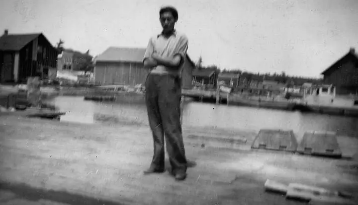 A man stands with folded arms on a dock beside a waterfront lined with wooden buildings and small boats.