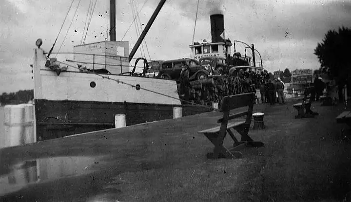 Black-and-white photograph of a vehicle-carrying vessel beside a pier, with cars, passengers, benches, and trees visible.