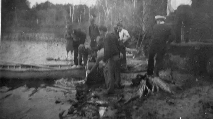 A group of people works beside a small boat at a wooded shoreline.