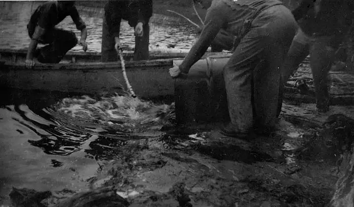 People work beside a wooden boat in shallow water, handling a large barrel as fish splash nearby.