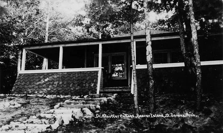 Black-and-white photograph of a small house with a broad porch among trees and rocks.