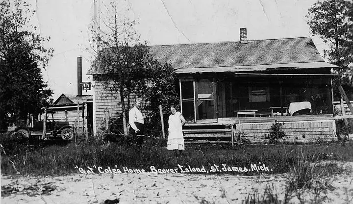 Black-and-white photograph of two people beside a shingled house, with a vintage automobile and handwritten caption.