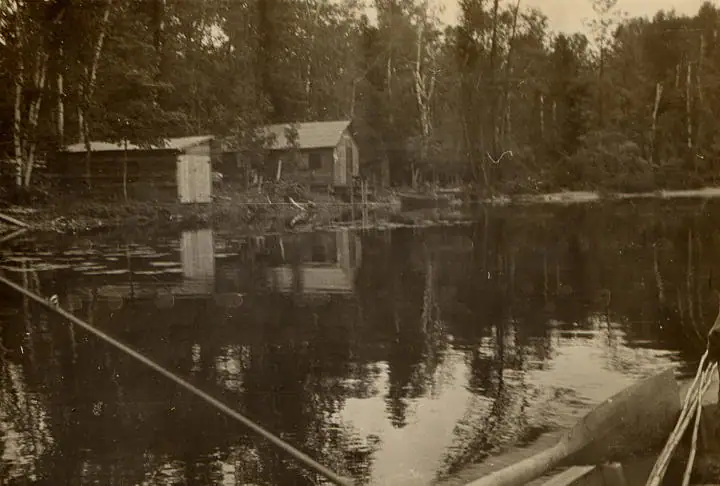 Sepia photograph of a wooded shoreline with a small cabin, its reflection, and part of a boat in the foreground.