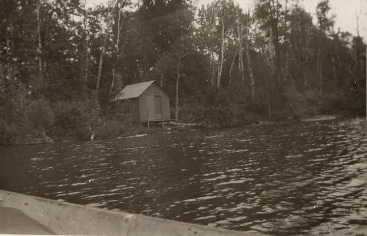 A small gabled shed stands at the wooded water's edge, with rippling water in the foreground.