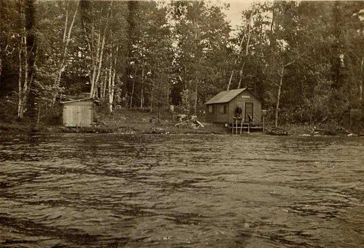 Sepia photograph of a small cabin and shed beside rippled water in a wooded setting.