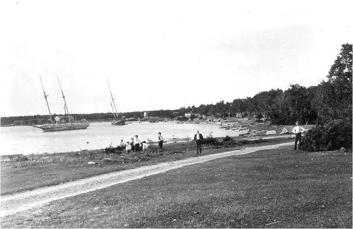 Black-and-white waterfront photograph with sailing vessels, people, a shoreline path, and wooded banks.