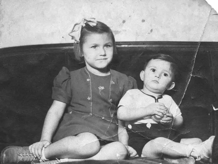 A girl and a toddler sit side by side on a cushioned bench in a black-and-white photograph.