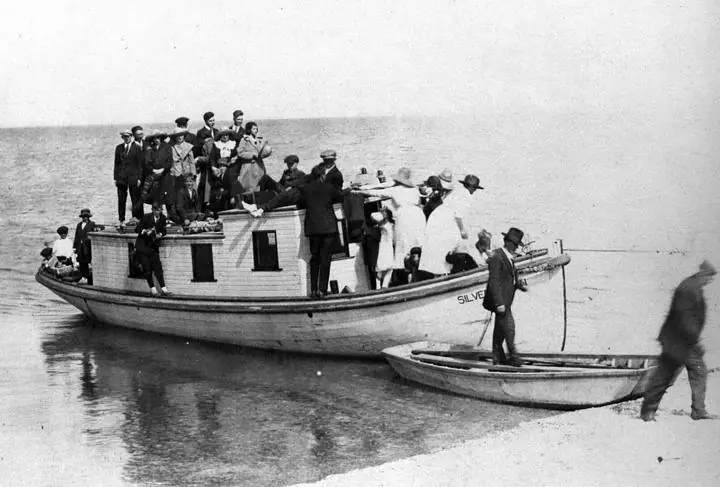 A small passenger boat crowded with people approaches a sandy shore beside a rowboat.