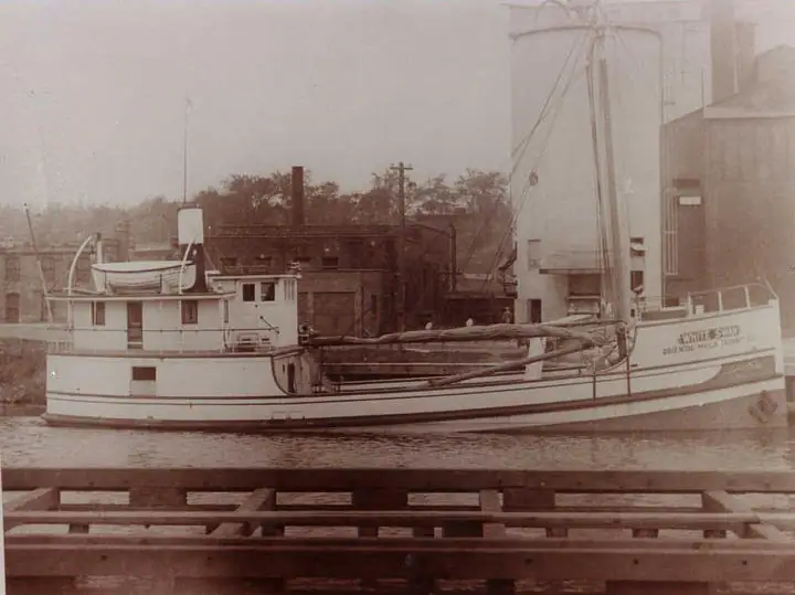 A white tugboat with hull lettering moored beside an industrial waterfront and wooden pier.