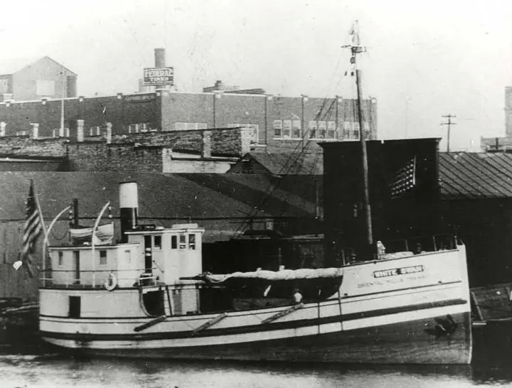 A tugboat with painted lettering is moored beside industrial buildings and a dock.