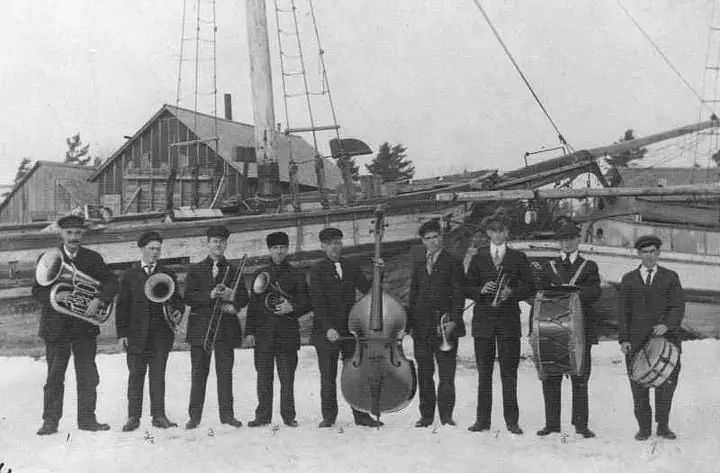 Nine musicians in dark suits pose with brass instruments, a double bass, and drums before a sailing vessel.
