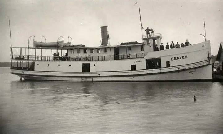 A white steamboat with a smokestack and people aboard moves across calm water.