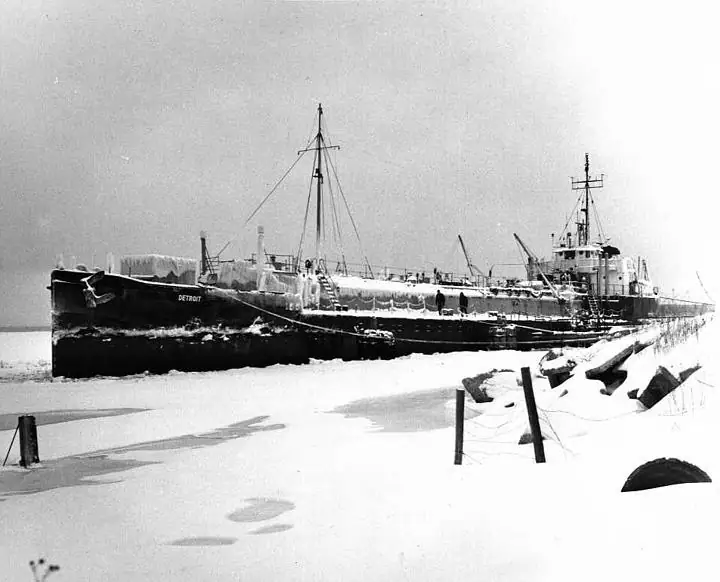 A large ship sits amid ice beside a snow-covered shoreline, with the hull name “DETROIT” visible.