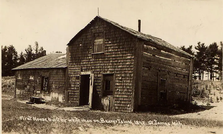 Sepia photograph of two weathered wooden buildings with shingled walls, open doors, and a handwritten caption along the bottom.