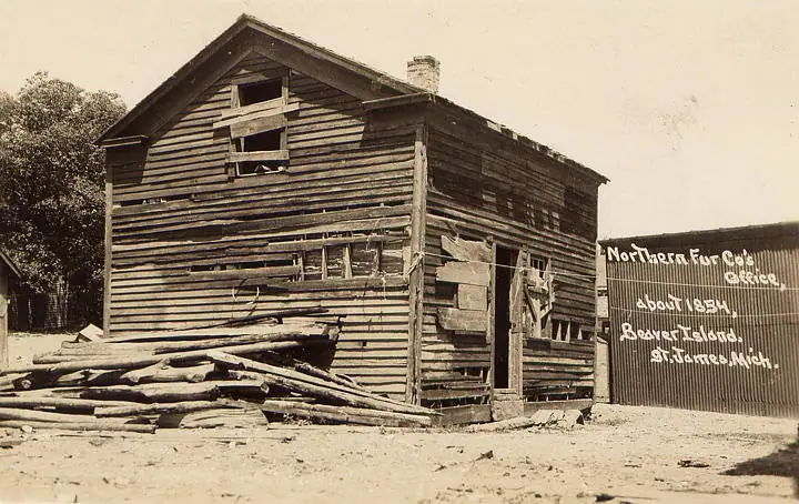 Dilapidated wooden office with a hand-lettered historical sign and a pile of logs in front.