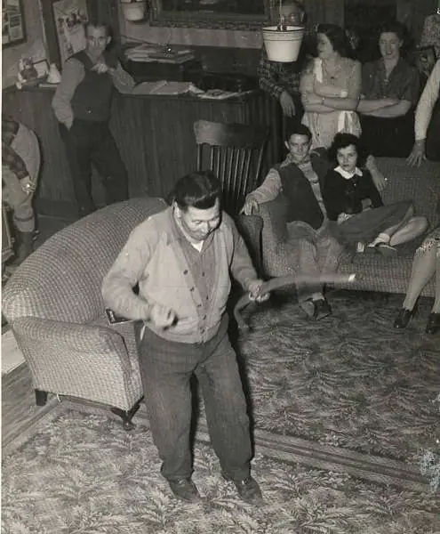 A man dances with a long object while a group watches from a furnished room.