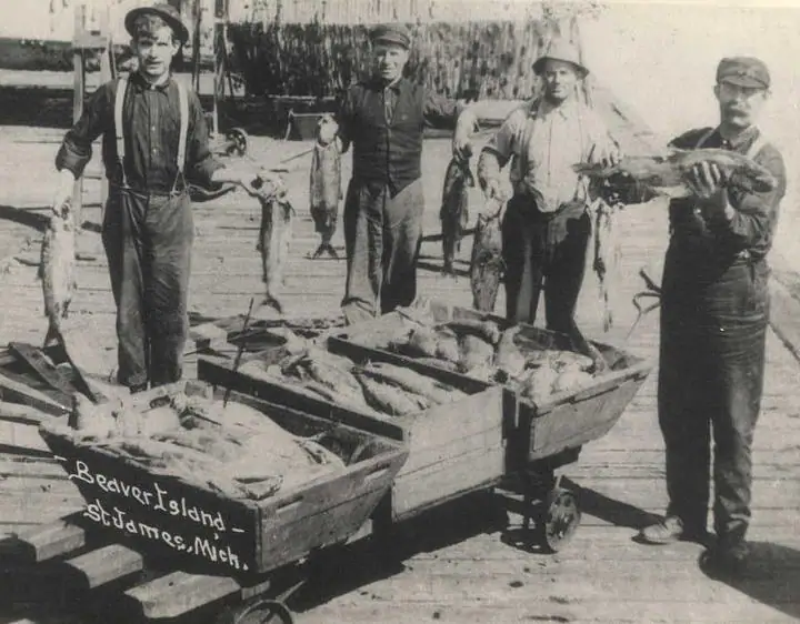 Four men display fish beside two wooden carts on a dock.