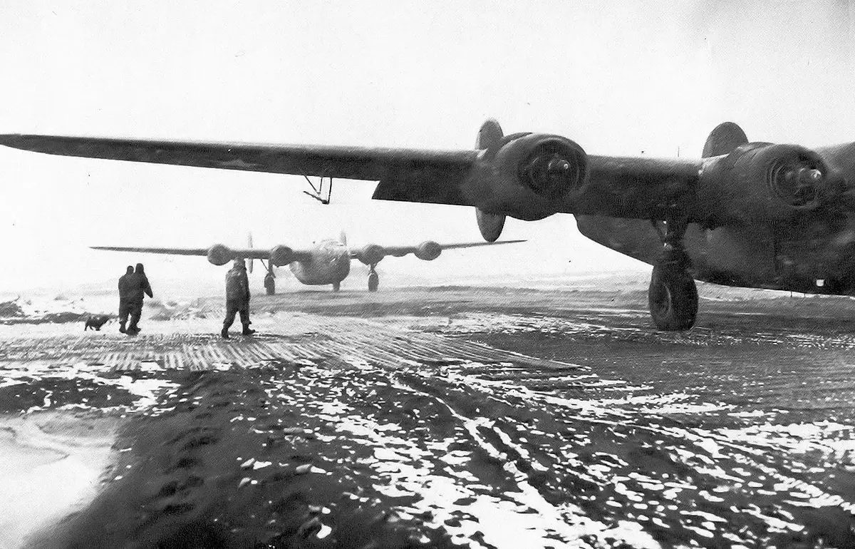 Two four-engine aircraft stand on a snow-covered airfield while people and a dog are nearby.