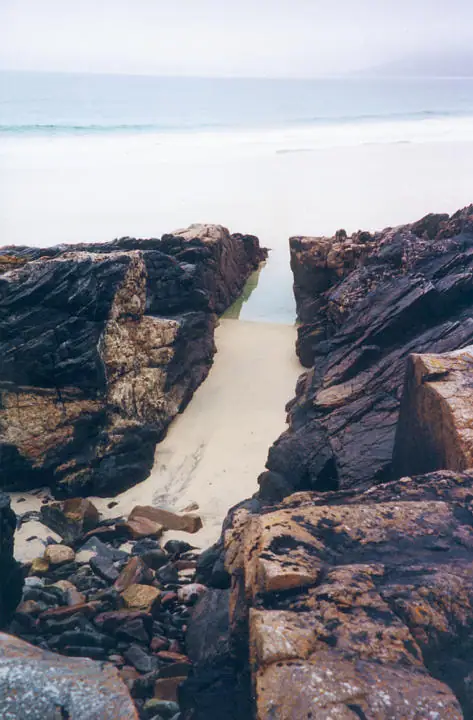 A sandy channel between dark layered rocks leads toward waves and a pale horizon.