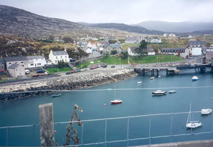 A harbor with small boats, waterfront houses, a road, and rocky hills beyond.