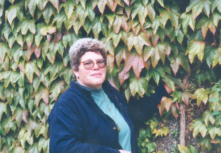A woman in glasses and a dark jacket poses beside a wall of leafy vines.