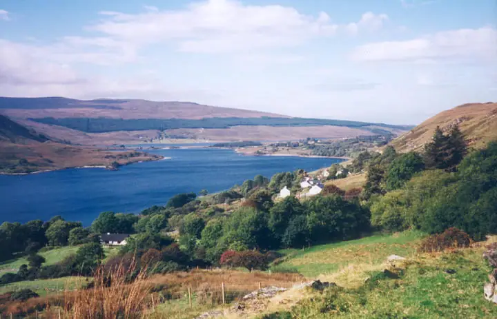 A broad blue body of water winds through rolling hills, with scattered houses and green fields in the foreground.