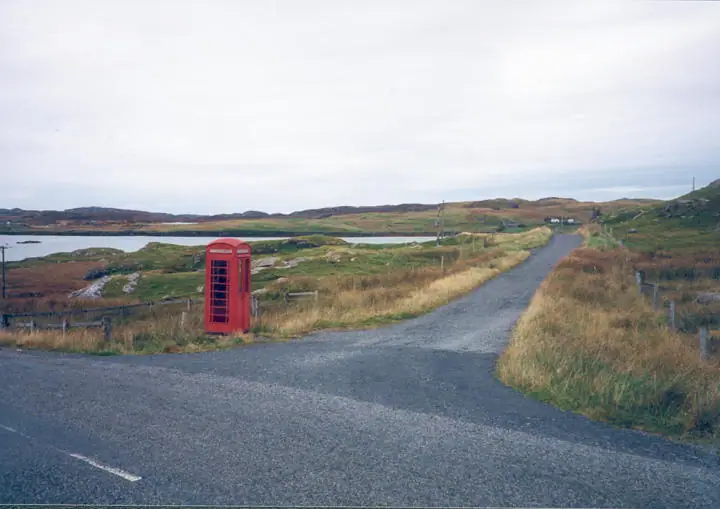 A red telephone box stands beside a rural road, with fenced grassland, water, and low hills beyond.